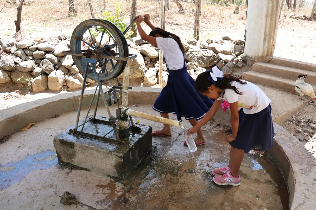 Ein Mädchen bedient einen Trinkwasserbrunnen, das andere fängt das geschöpfte Wasser auf.